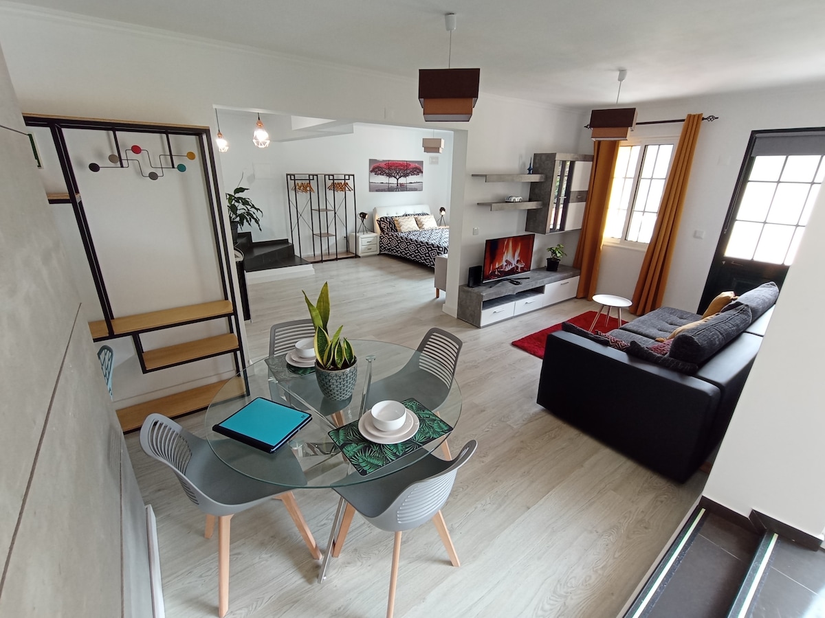 An open living area is presented, featuring a modern glass dining table surrounded by sleek chairs. A comfortable sofa sits nearby, alongside a media unit with a decorative fireplace. Large windows allow natural light to flood the space, highlighting the contemporary decor.