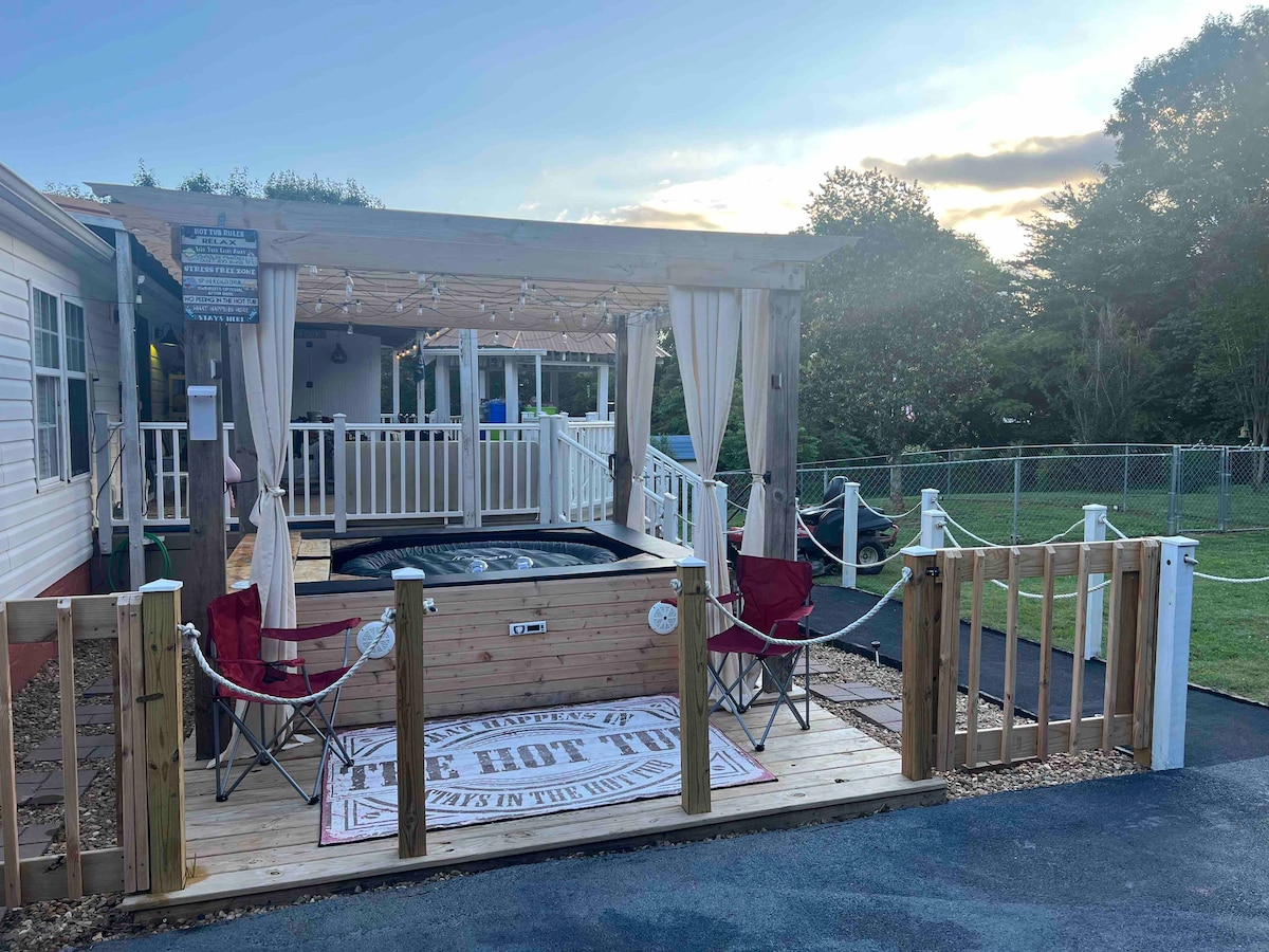 A hot tub area is surrounded by wooden fencing and features two red chairs, a patterned mat, and curtains for privacy. The rustic structure is complemented by a covered deck nearby, providing space for relaxation under the sky.