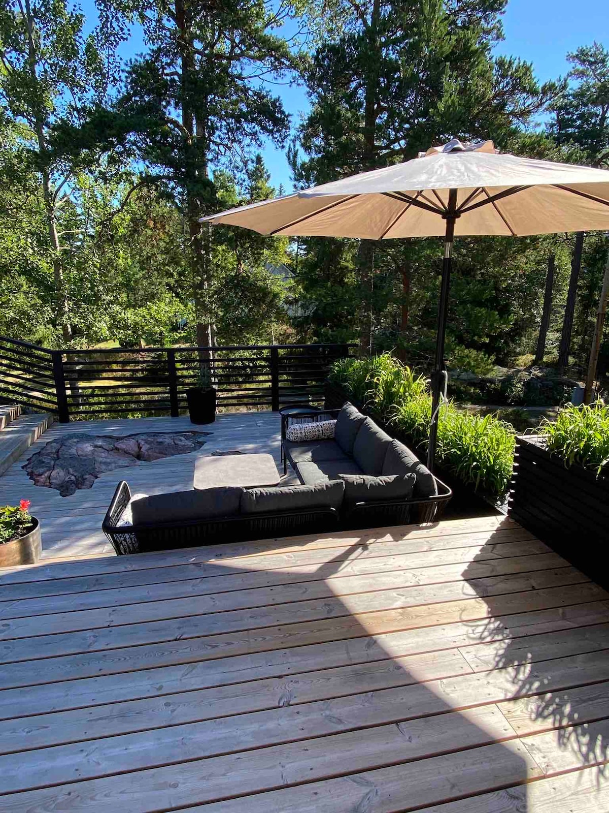 A shaded lounge area is depicted with a sectional sofa arranged on a wooden deck, surrounded by lush greenery. An umbrella provides coverage, while planters add a touch of nature. The tranquil setting is designed for relaxation and comfort amid an outdoor environment.