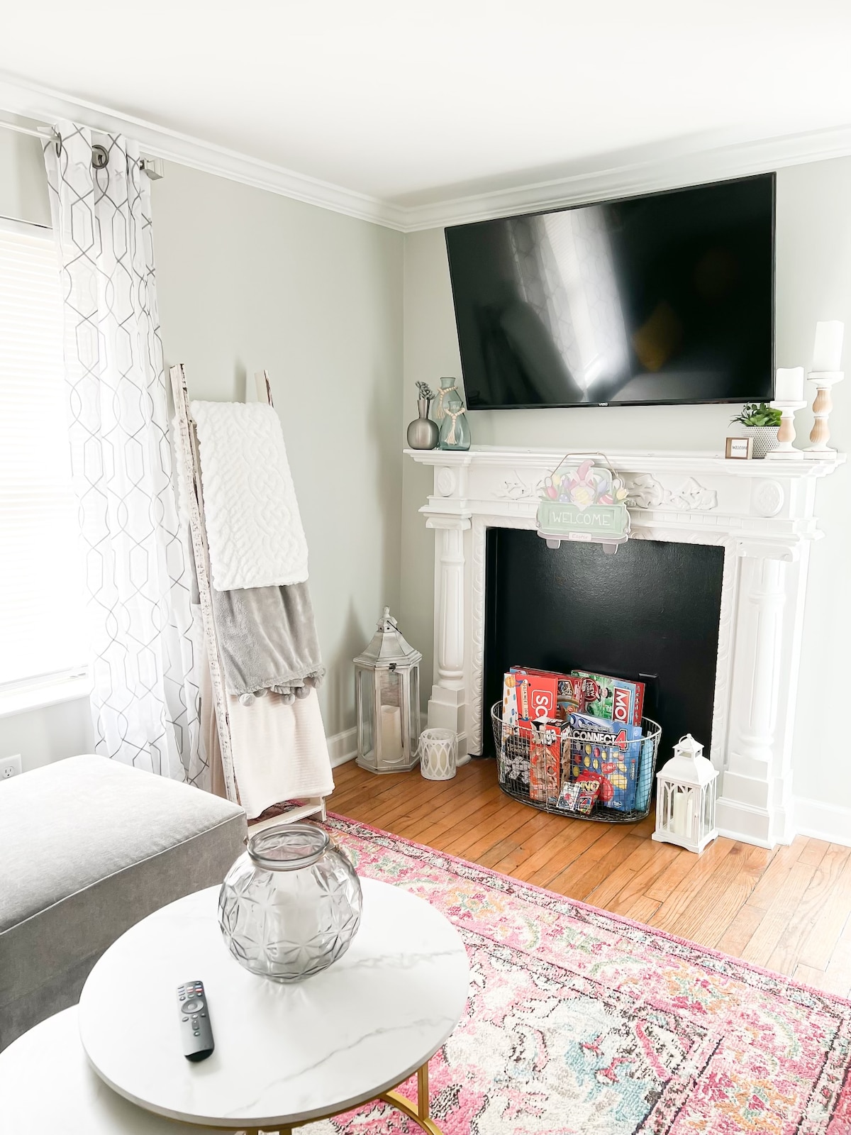 A cozy living area features a decorative fireplace with a modern television mounted above. Light-colored curtains frame a window, and a stylish rug adds warmth to the hardwood floor. A glass table with a vase sits in front of a comfortable couch, creating an inviting space.