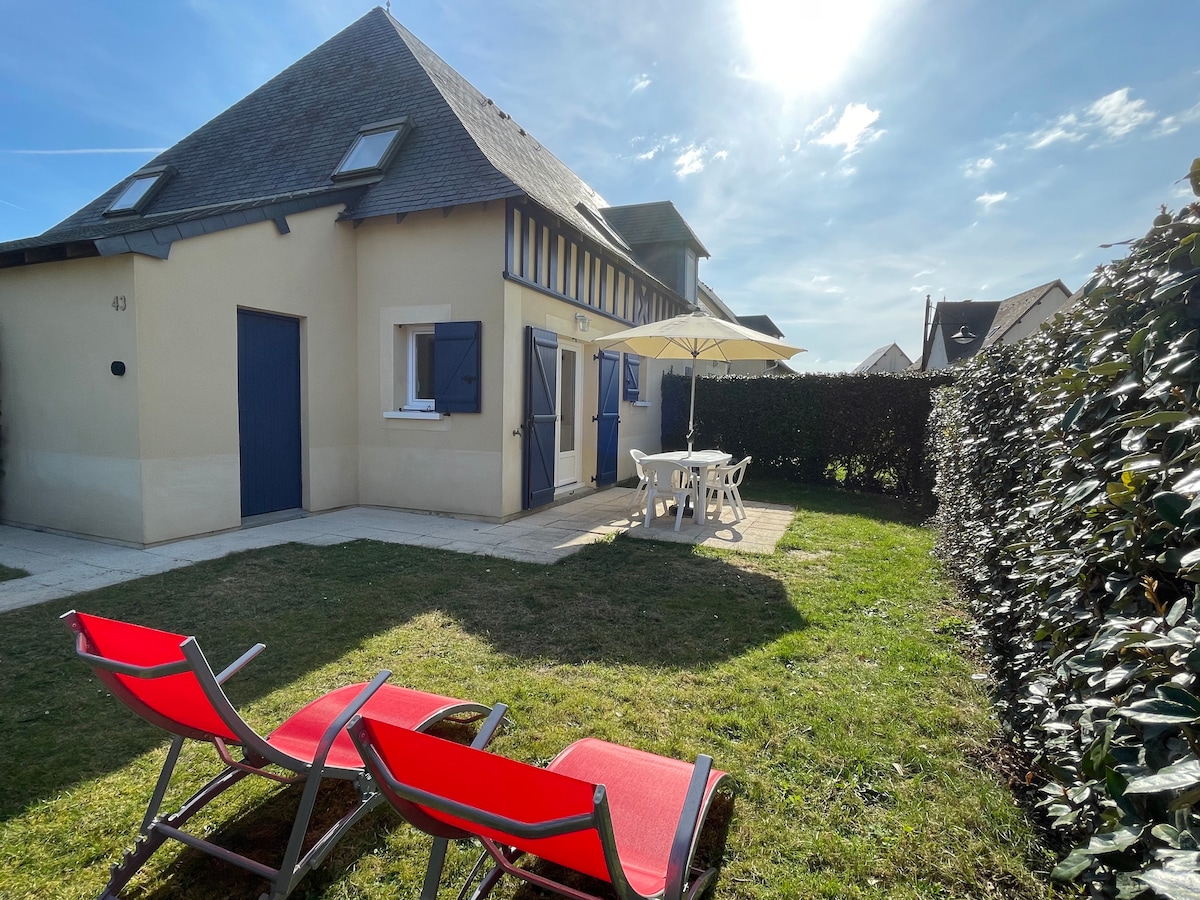 The exterior of the house is presented with a well-maintained garden featuring two red lounge chairs. A dining table with chairs sits under an umbrella, providing a shaded area. The sunlight illuminates the space, highlighting the house's architectural details.