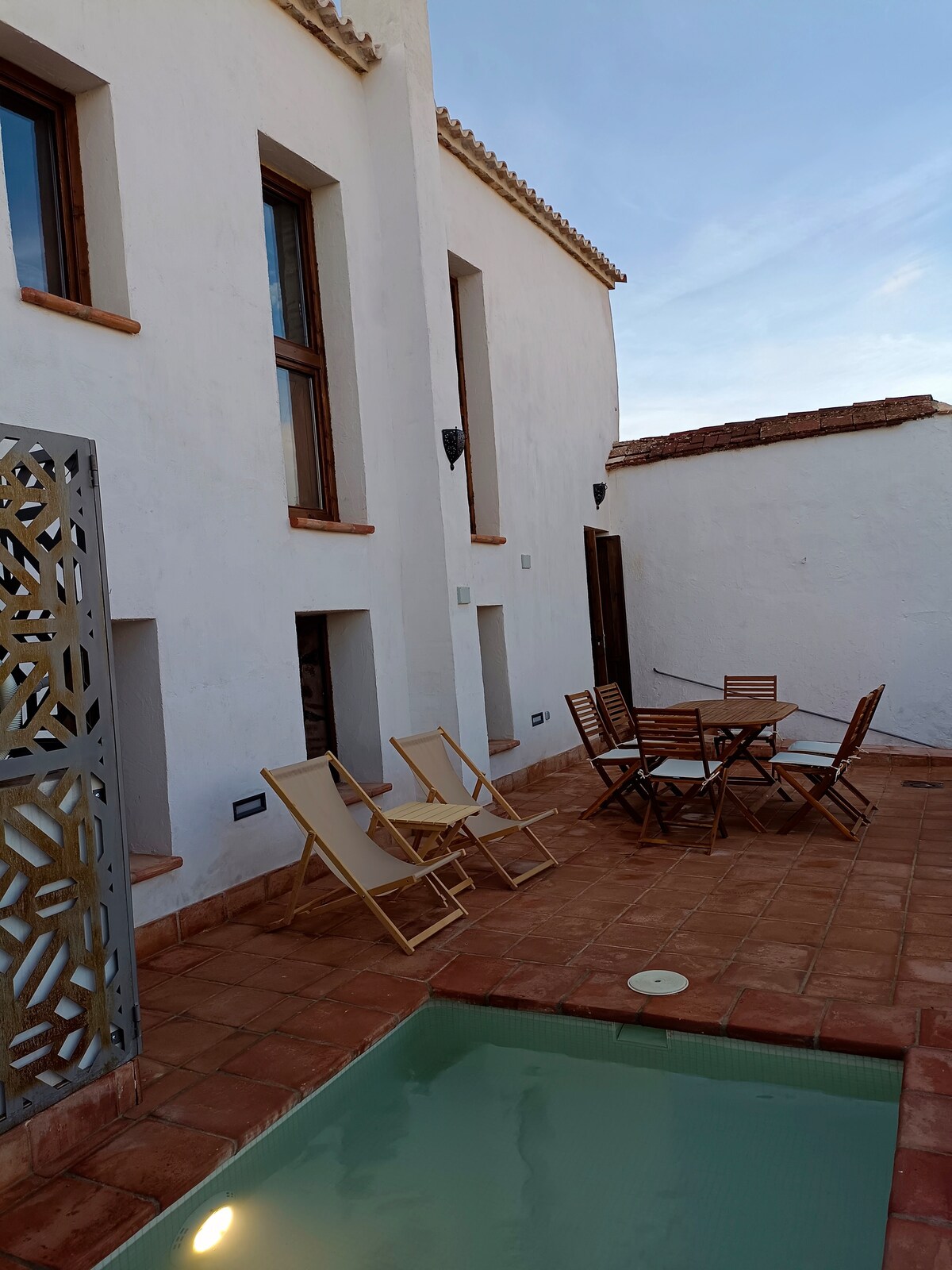 An outdoor terrace features inviting seating arrangements, including a wooden dining table surrounded by folding chairs. A small pool is visible, complemented by a textured partition. Tall windows illuminate the white exterior walls, reflecting a blend of rustic charm and modern design.