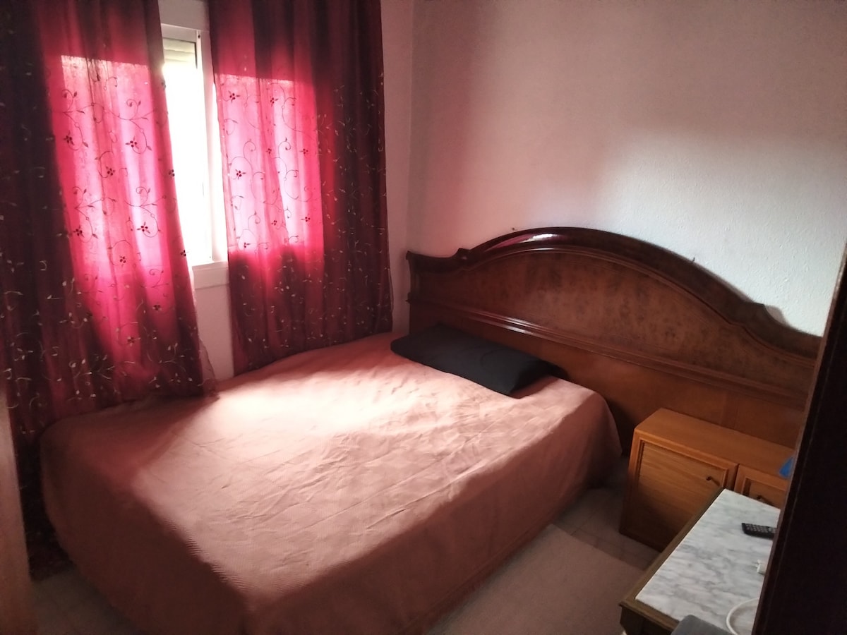 A cozy sleeping area features a wooden headboard and a soft bedspread in neutral tones. Light filters through a window dressed with maroon curtains, creating a warm ambiance. A small bedside table is positioned next to the bed, adding to the room's functionality.