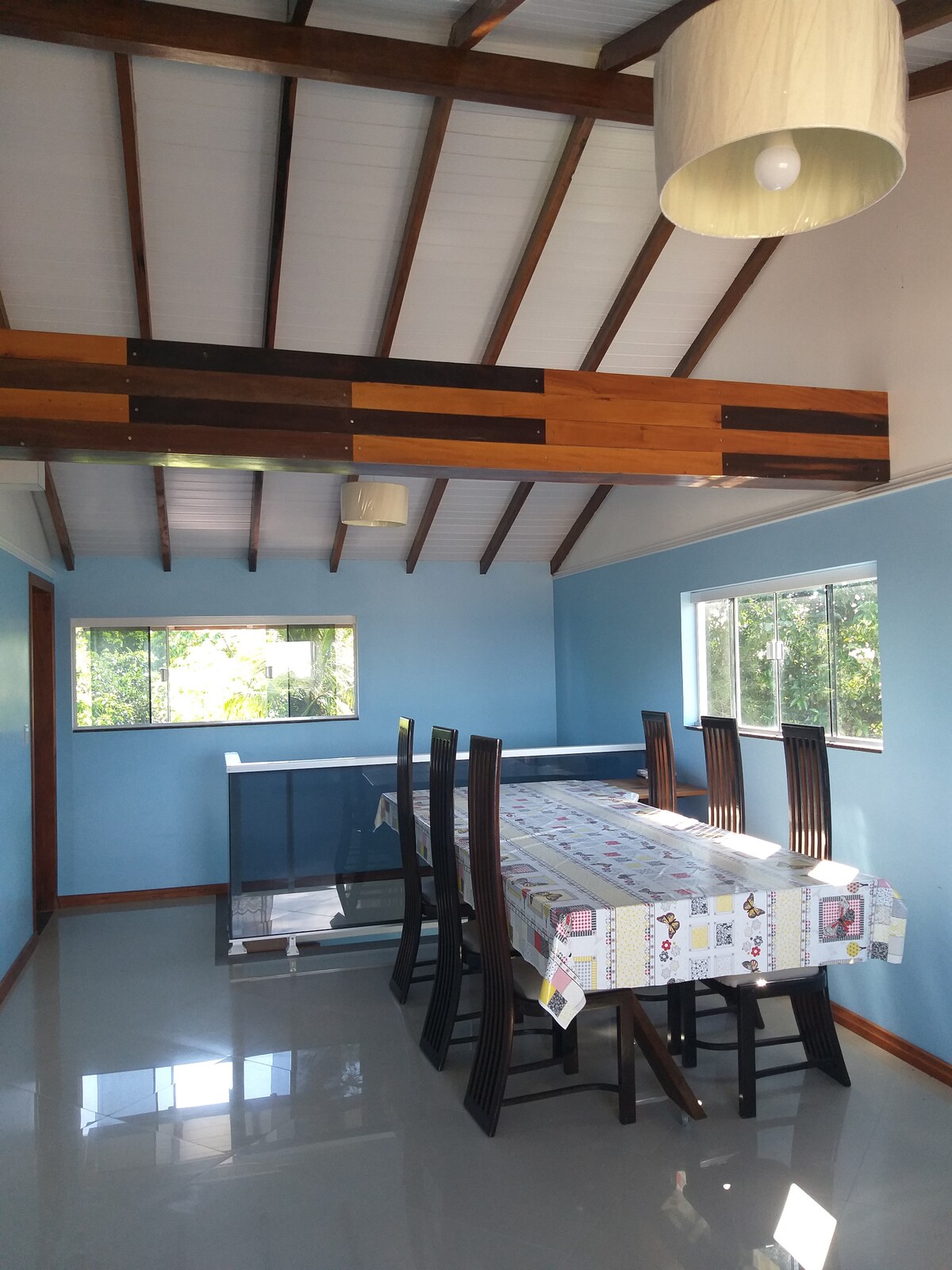 A dining area features a large table surrounded by dark wooden chairs, suitable for eight. Natural light enters through multiple windows, highlighting the light blue walls and polished floor. The ceiling showcases exposed beams, enhancing the spaciousness of the room.