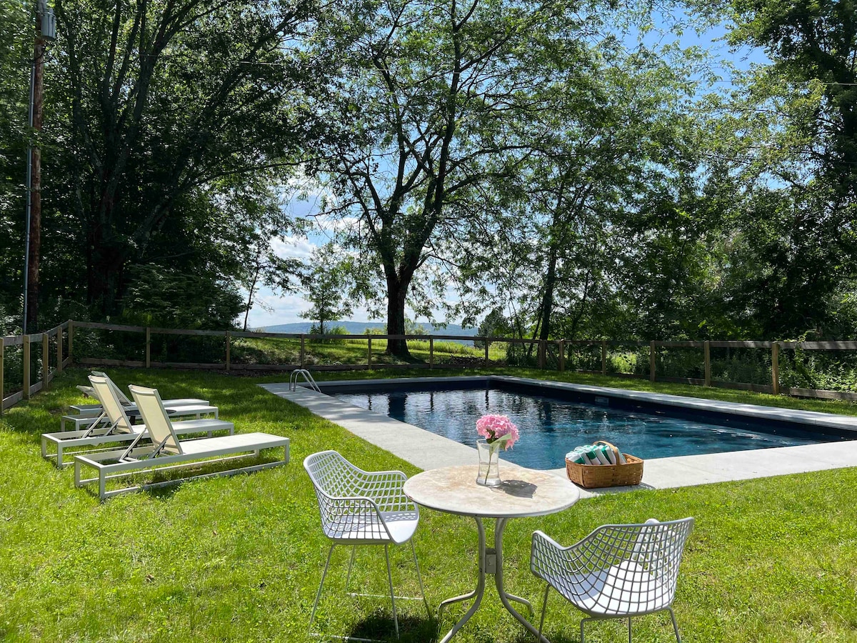A serene outdoor space features a swimming pool surrounded by a lush green lawn. Several lounge chairs are positioned nearby. A table set with a floral centerpiece and a basket is situated on the grass, providing a relaxing atmosphere under a clear blue sky.