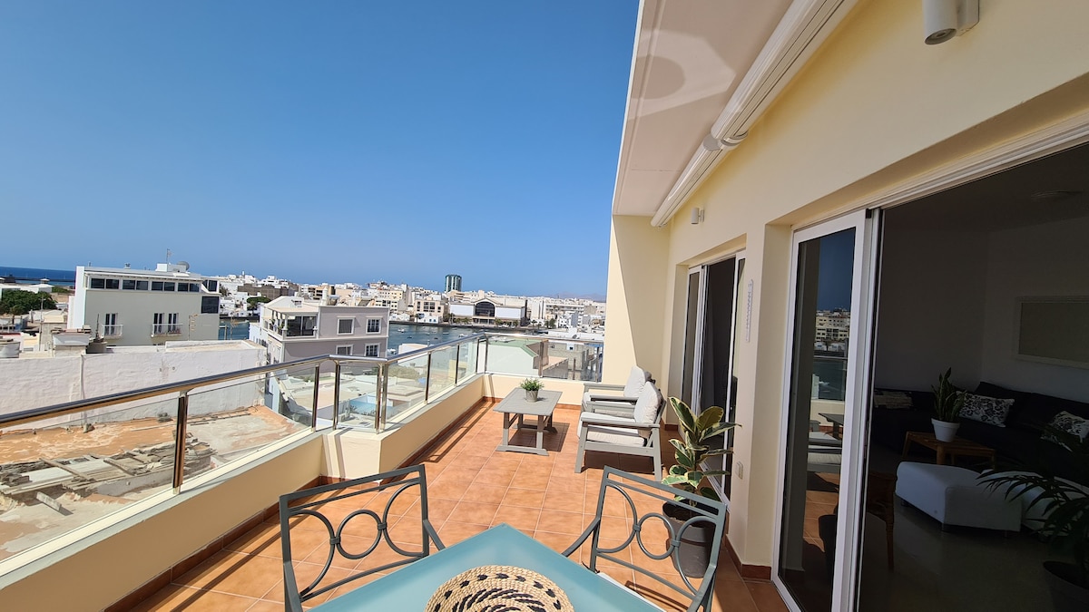 A spacious terrace is visible, featuring outdoor seating and a dining table. The area offers views of the cityscape and the ocean in the distance. Large glass doors provide access to the interior, allowing natural light to brighten the space.