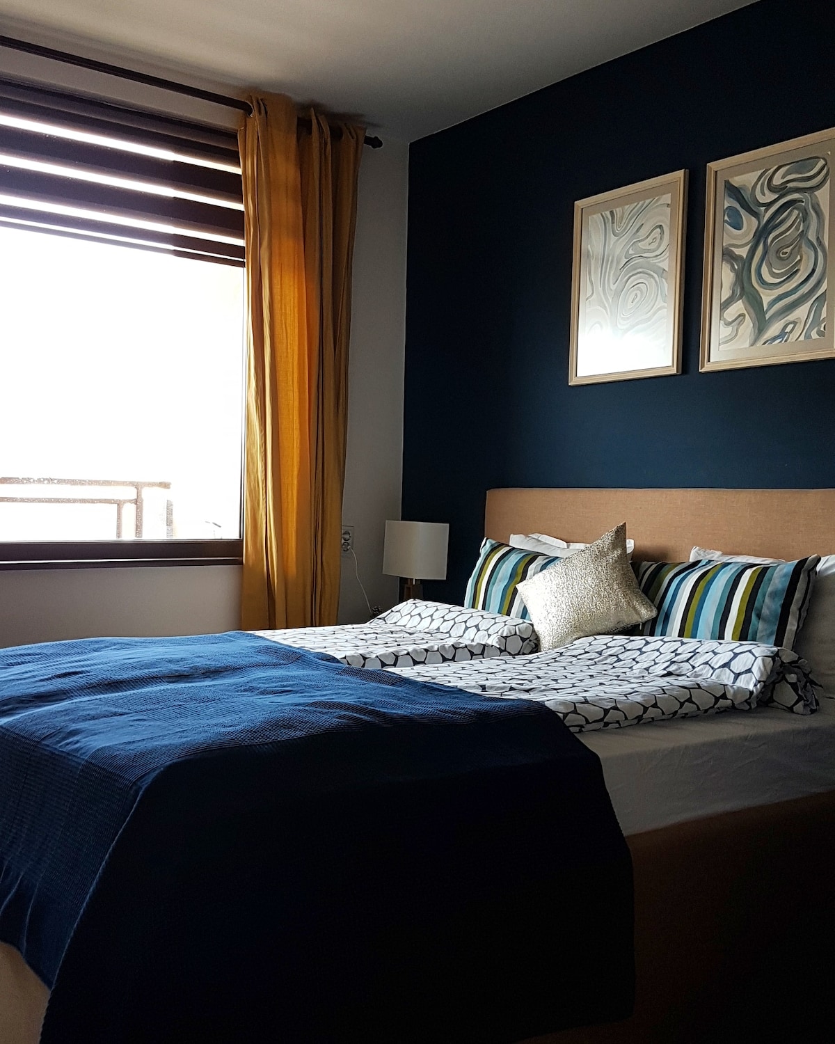The bedroom features a spacious bed adorned with patterned bedding and a decorative pillow. Warm yellow curtains frame a large window, allowing natural light to fill the room. Two pieces of abstract wall art hang above the bed, complementing the deep blue accent wall.
