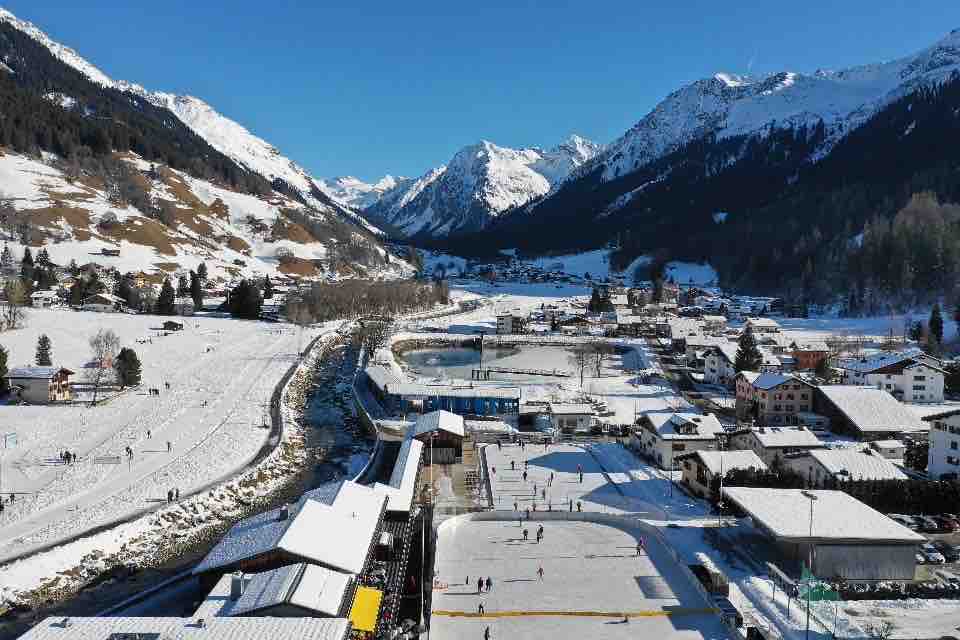 A panoramic view captures a snowy village surrounded by mountains. An ice-skating rink is visible, with people enjoying the winter landscape. The river flows gently beside the area, and the clear blue sky complements the serene atmosphere.