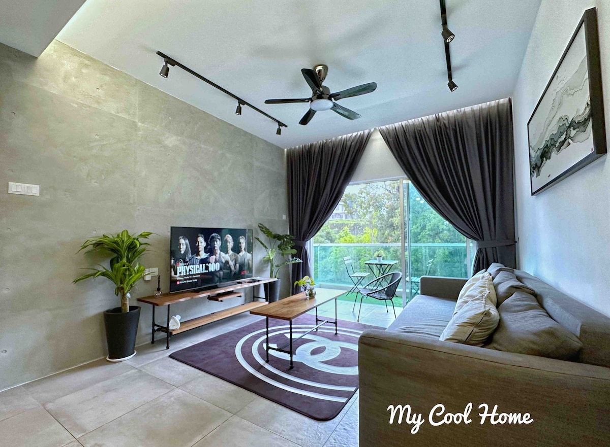 The living area is furnished with a three-seater sofa bed and a coffee table, complemented by a flat-screen smart TV. Large windows allow natural light to fill the space, with views of greenery visible outside. Two lounge chairs are positioned next to the windows.