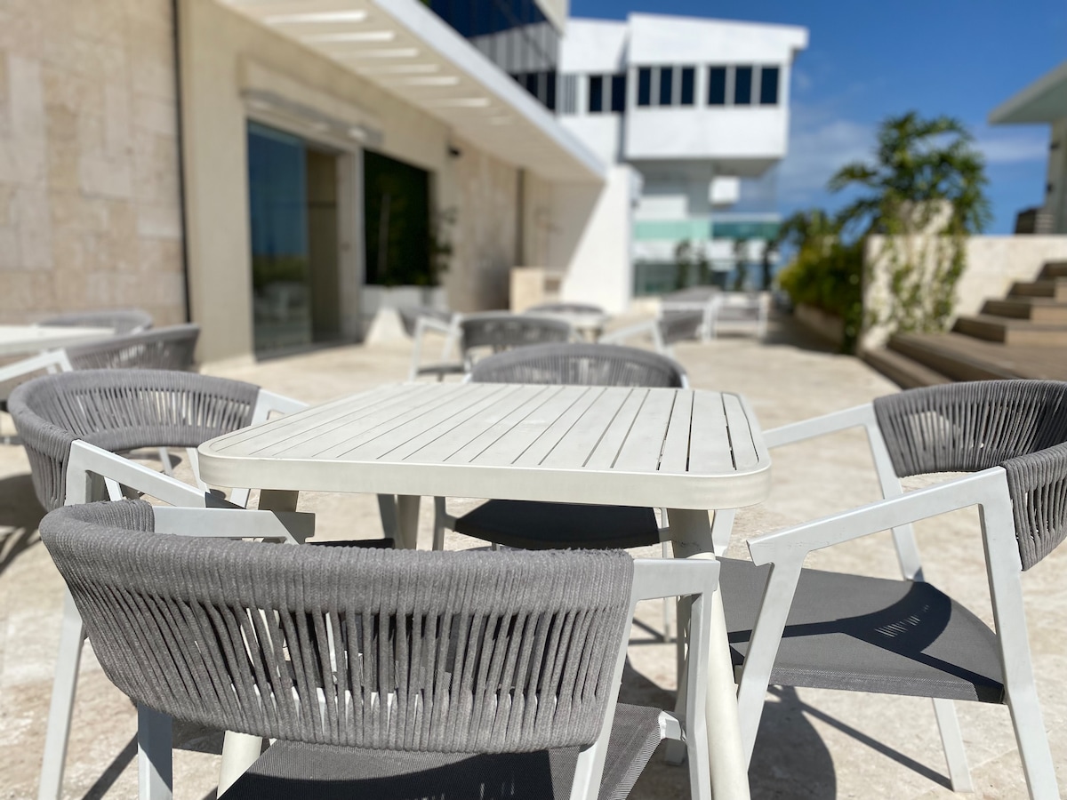 An outdoor dining area features a rectangular table surrounded by six round chairs. The chairs are designed with gray woven seats and backs. Sunlight brightens the space, highlighting the clean lines of the furniture against a backdrop of modern architecture.