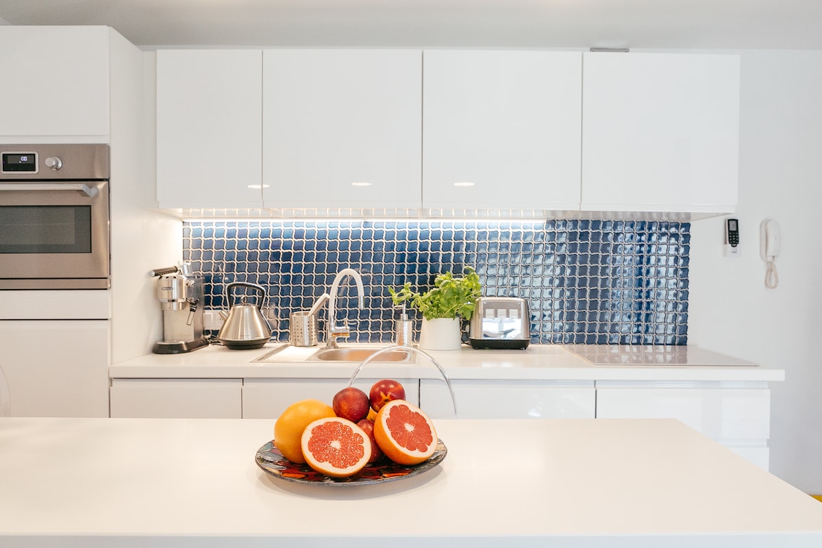 A modern kitchen is presented with white cabinetry and a blue tiled backsplash. A bowl of fresh fruit, including grapefruit and apples, is centered on a sleek island countertop. Stainless steel appliances, including a kettle and toaster, are visible, contributing to a functional cooking space.