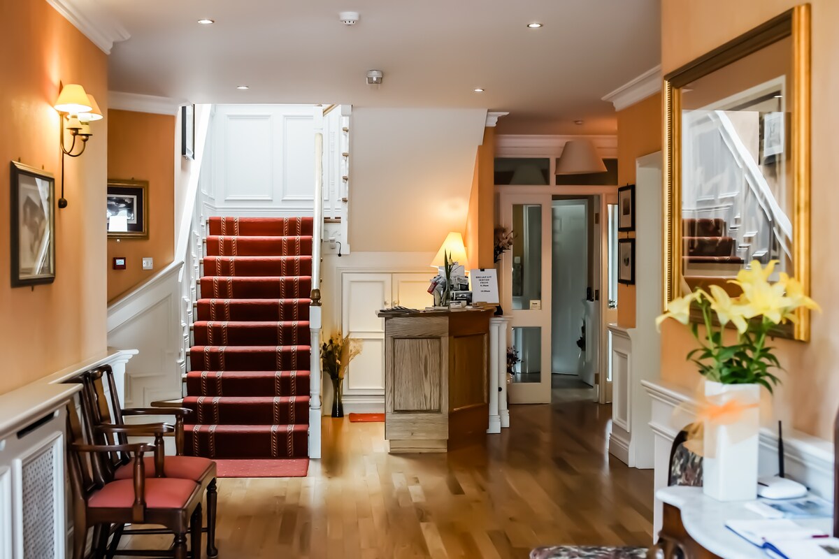 A welcoming lobby area is presented with a wooden reception desk situated to the left. The space features warm-toned walls, polished wooden floors, and a staircase with a red carpet. Two wooden chairs are placed against the wall, adding to the inviting atmosphere.