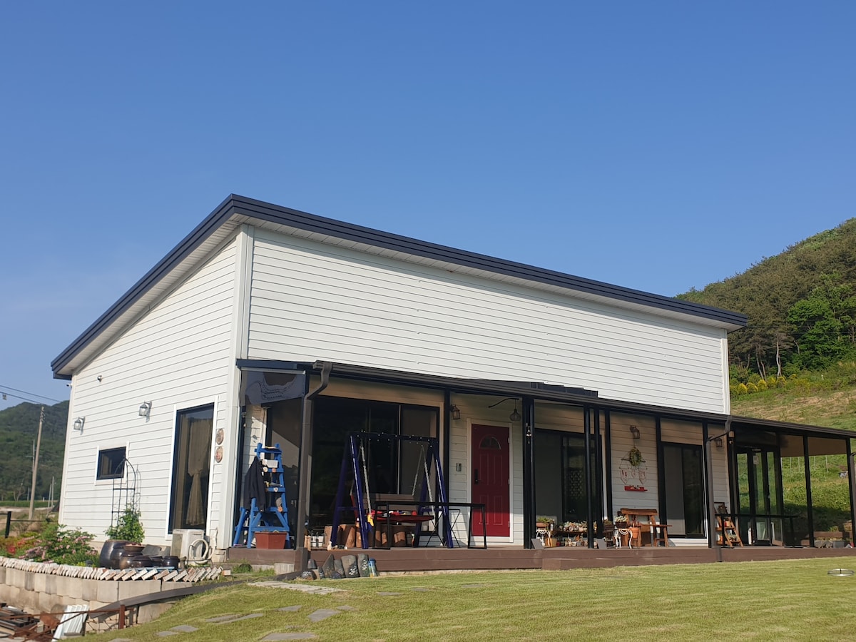 A spacious farmhouse with a modern exterior is depicted, featuring white siding and a contrasting red door. Large glass windows provide unobstructed views of the surrounding landscape. The well-maintained yard includes a broad grassy area, creating an inviting outdoor space.