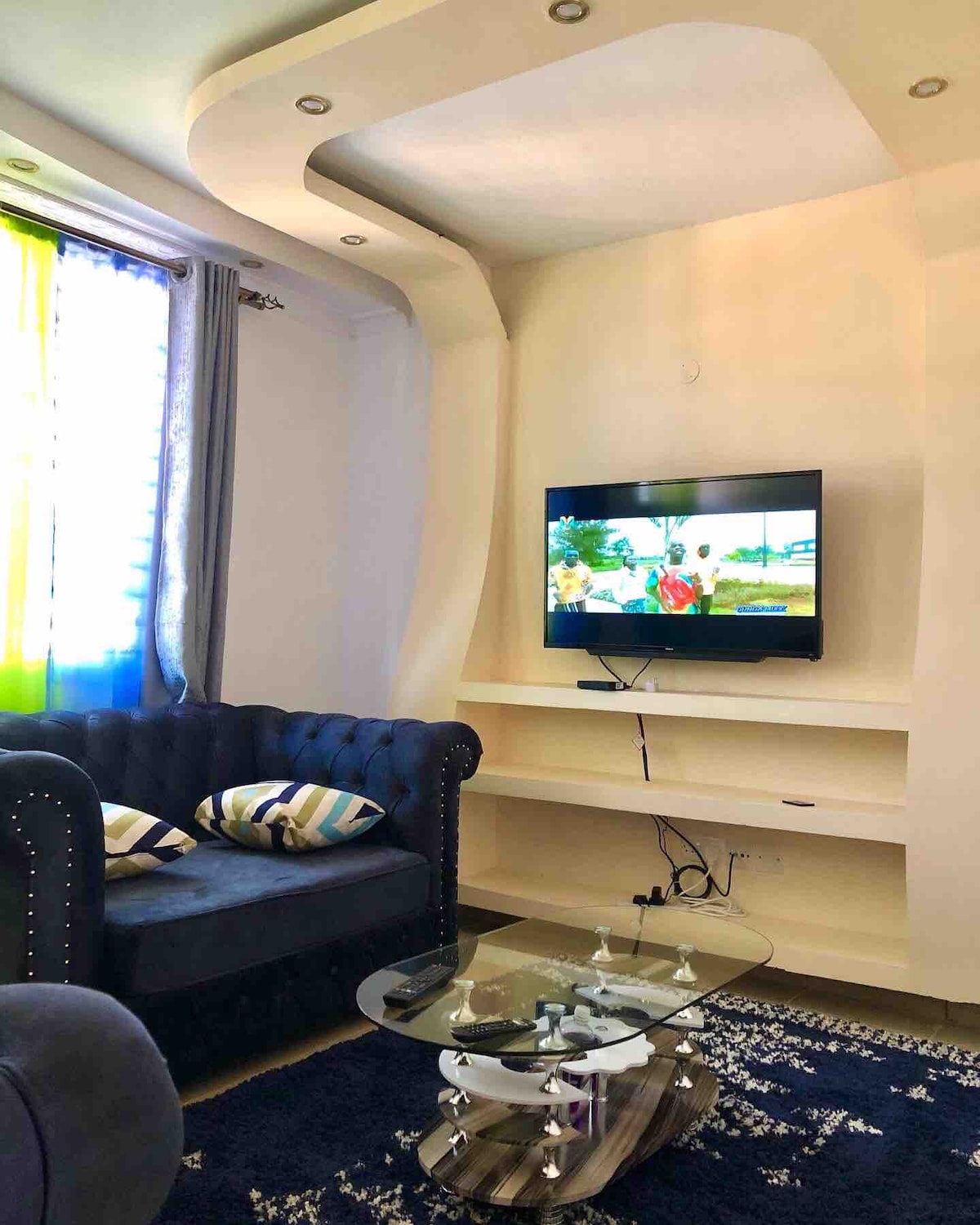 A living area features a comfortable blue sofa with decorative pillows, positioned opposite a flat-screen television mounted on a light-colored wall. A glass coffee table rests on a vibrant area rug, with natural light streaming in from curtains that blend shades of blue and green.