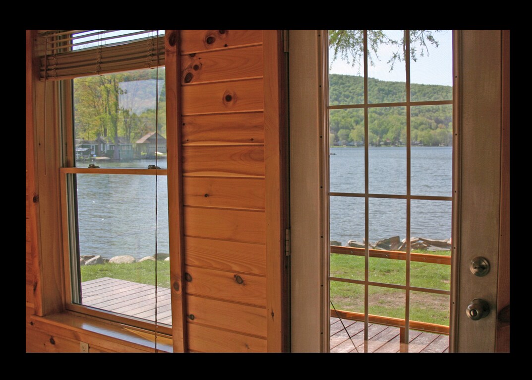 A view from inside the cottage reveals large windows framed by warm wooden paneling. The door opens to a deck overlooking DeRuyter Lake and lush green hills in the distance. Natural light fills the space, emphasizing the serene lakeside atmosphere.