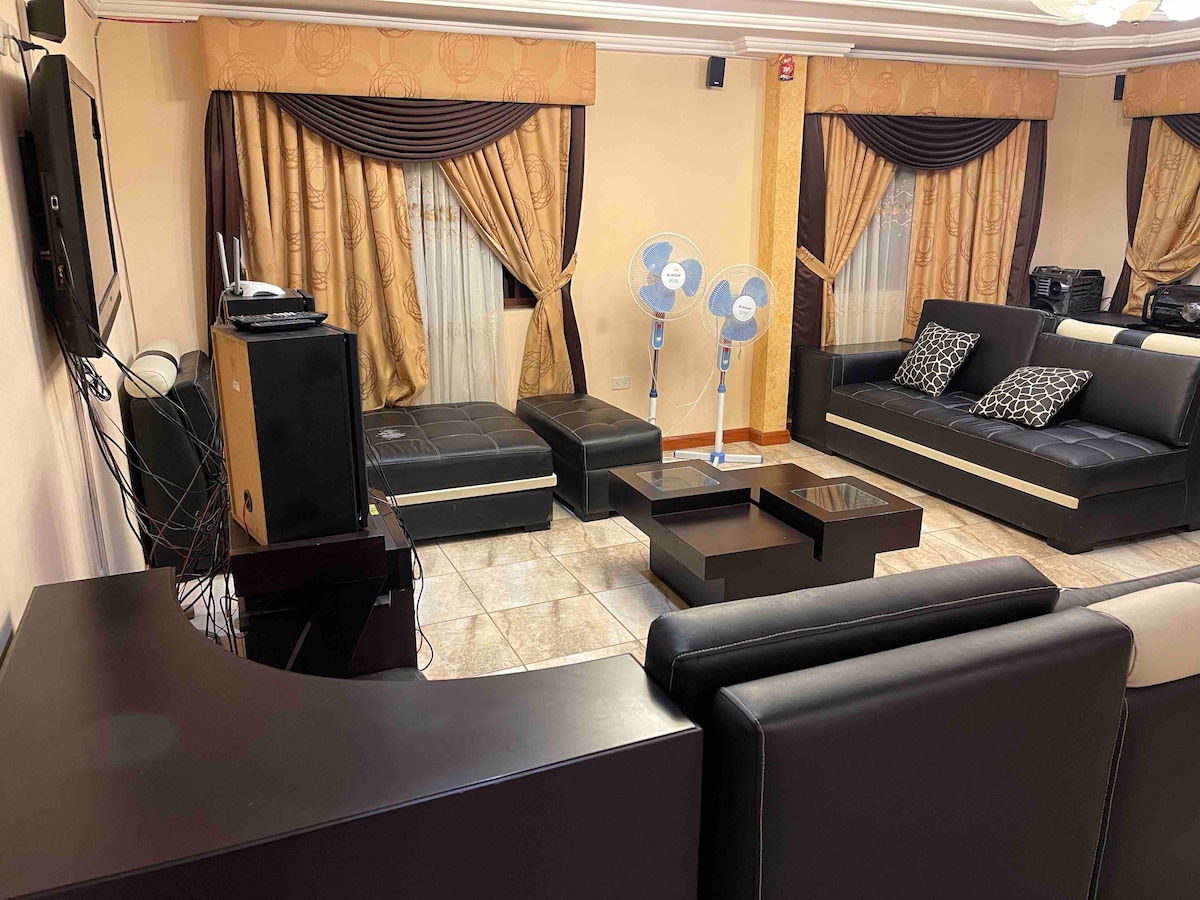 A spacious living area features dark leather seating complemented by elegant cushions. Two standing fans are positioned near large windows dressed with golden curtains. A sleek coffee table sits at the center, surrounded by a warm, tiled floor.
