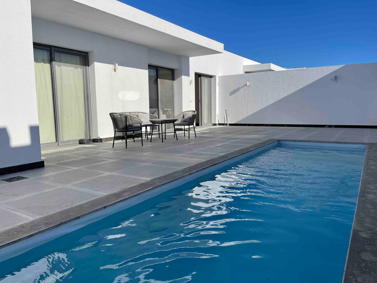 A private heated pool is framed by a spacious patio featuring a small seating area with two chairs and a table. Large sliding glass doors provide access to the interior, illuminated by natural light. The clean lines of the architecture contrast with the clear blue sky.
