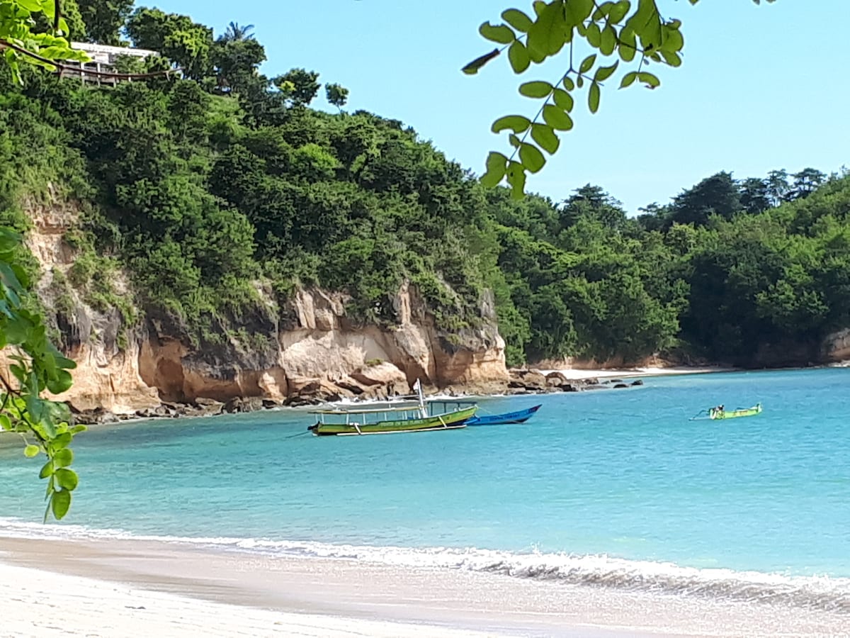 A serene coastal view showcases a calm turquoise sea framed by lush greenery and cliffs. Two boats float gently on the water near a sandy beach, inviting relaxation and exploration. The clear sky enhances the peaceful atmosphere of the Ekas beach area.
