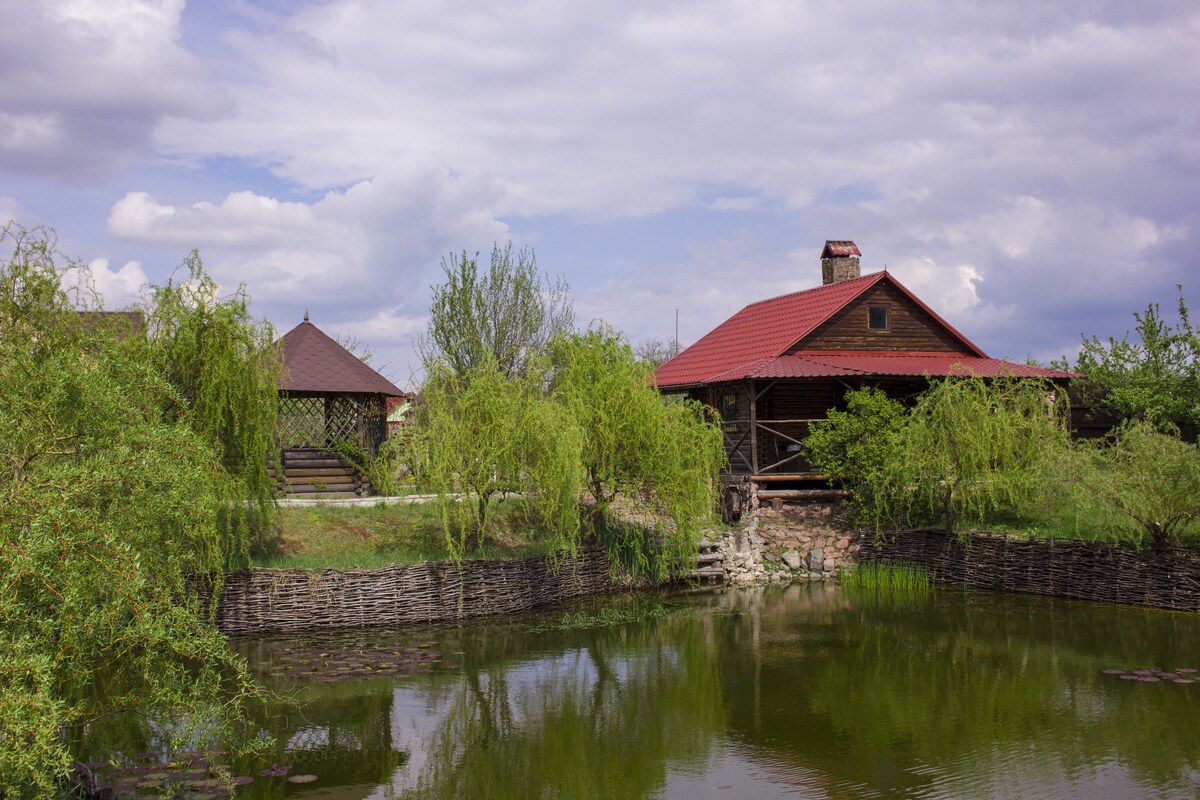 A charming hunting cottage is situated near a tranquil pond, framed by lush greenery and round willows. The rustic structure features a red roof and wooden details, while a gazebo stands nearby. Scenic views of the landscaped area invite relaxation and nature appreciation.