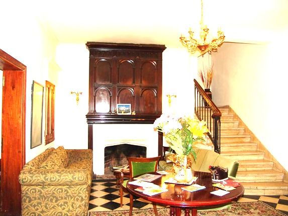 A warmly decorated living area showcases a patterned green sofa and a beige couch arranged around a round table. A large wooden fireplace is set against a wall adorned with dark wood paneling. A chandelier hangs above, and a staircase is visible in the background.