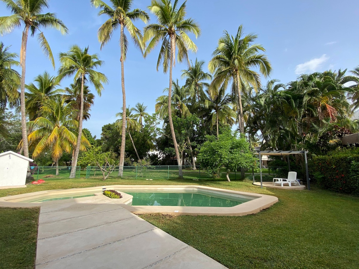 A private pool is surrounded by lush greenery and palm trees, creating a peaceful outdoor space. A concrete pathway leads to the pool area, where comfortable lounge chairs are positioned nearby, inviting relaxation under the sun.