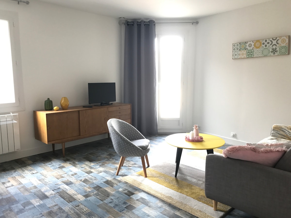 A bright living area features a gray sofa and a round yellow coffee table, creating an inviting space. A mid-century wooden console holds a television. Light filters through the large windows, accompanied by gray curtains. Colorful wall art adds a touch of creativity to the neutral palette.