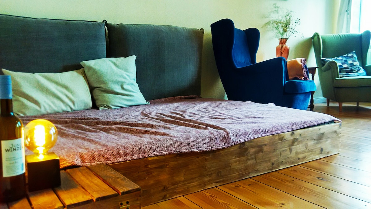 A spacious lounge area is highlighted by a low wooden platform bed covered with a soft blanket and several pillows. A stylish blue armchair and a green chair add seating options, while a small table holds a bottle of wine and a warm-lit lamp, creating a cozy atmosphere.