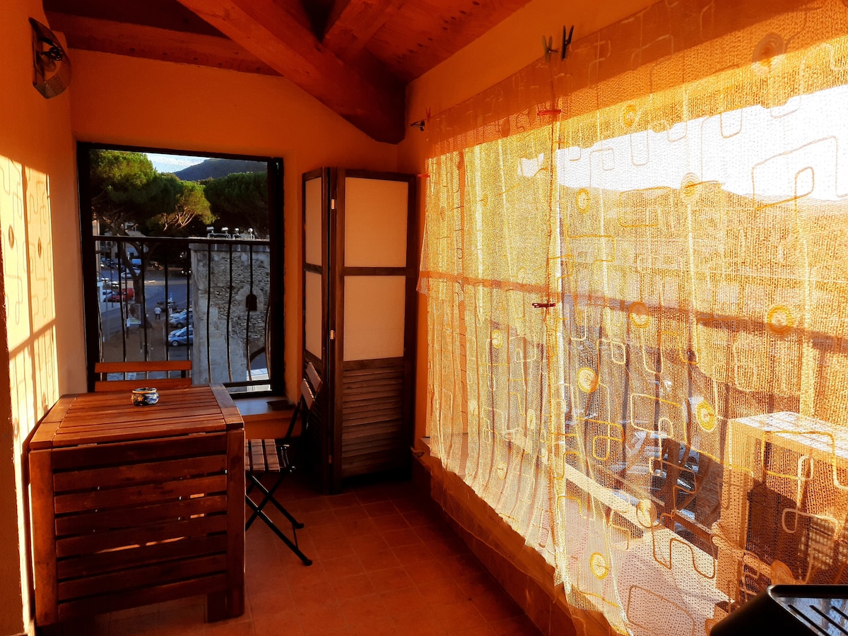 A small terrace features a wooden table and two folding chairs, illuminated by natural light. Sheer curtains create a soft barrier, allowing views of the surrounding landscape. Wooden beams highlight the ceiling, enhancing the cozy atmosphere of the outdoor space.