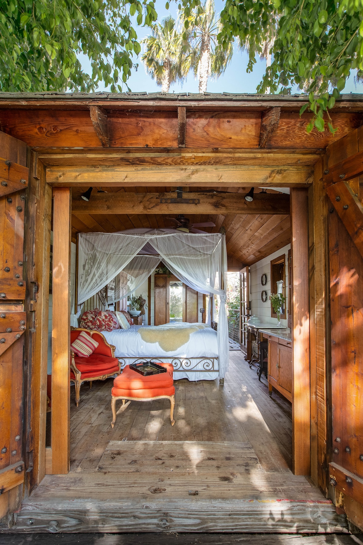 A welcoming cottage entrance reveals a spacious sleeping area adorned with a canopy bed draped in sheer fabric. Colorful pillows are arranged on the bed, while a bright orange chair and wooden furnishings bring warmth to the space. Natural light filters in from large windows, enhancing the rustic wooden interior.
