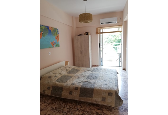 A spacious bedroom features a large bed with a patterned quilt. A simple closet is visible against the wall, and a vibrant piece of wall art adds a touch of color. Natural light filters in through glass doors that lead to a small balcony.