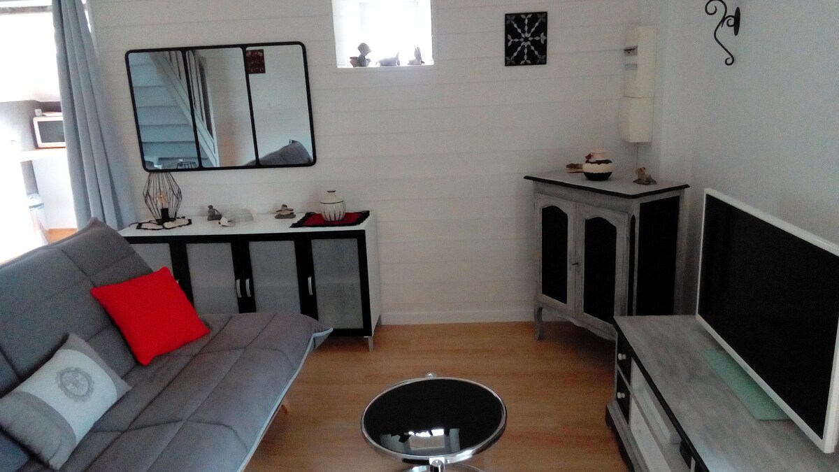 The living area features a modern gray sofa with a red accent pillow, complemented by a circular glass coffee table. A large mirror and decorative wall clock are mounted above a sleek cabinet, while a minimalist TV stand is placed against the wall, creating a cohesive space.