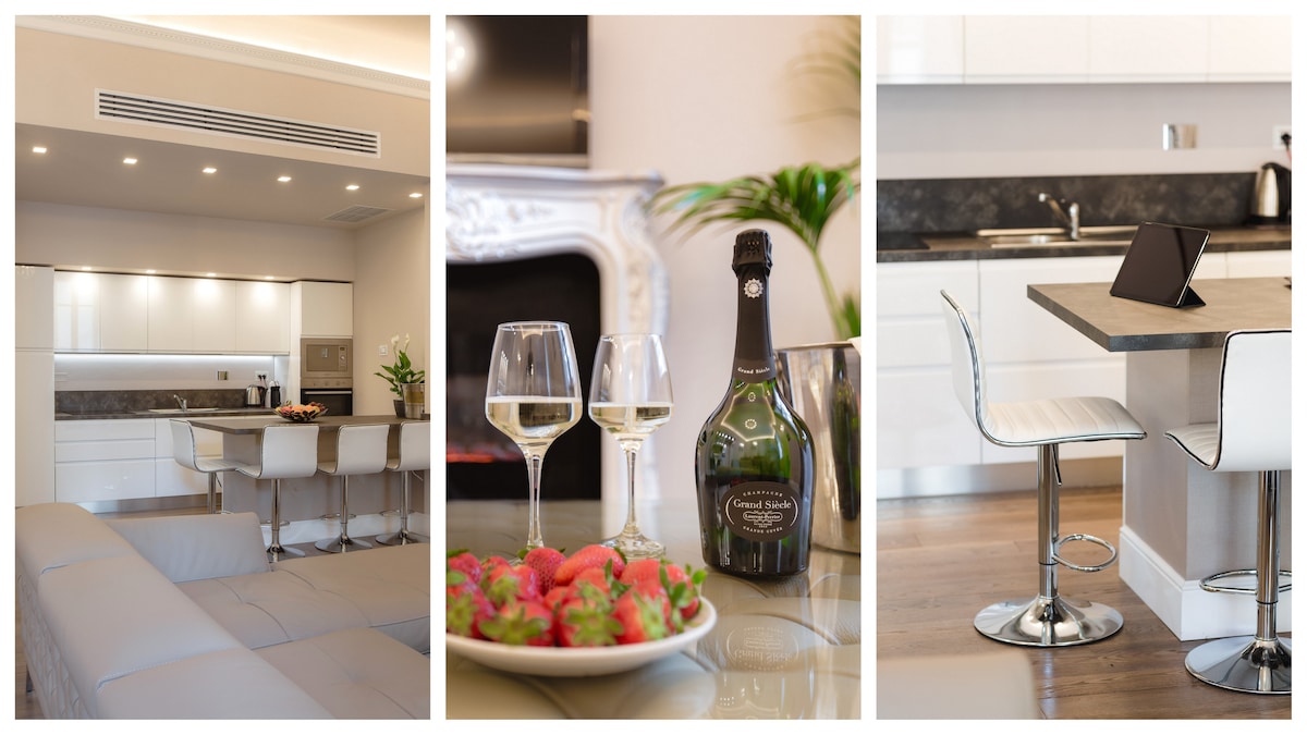 A modern kitchen and dining area is showcased, featuring sleek white cabinetry and a spacious island bar with high stools. Glasses of sparkling wine are set beside a bottle, accompanied by a plate of fresh strawberries, inviting a sense of leisure and elegance.
