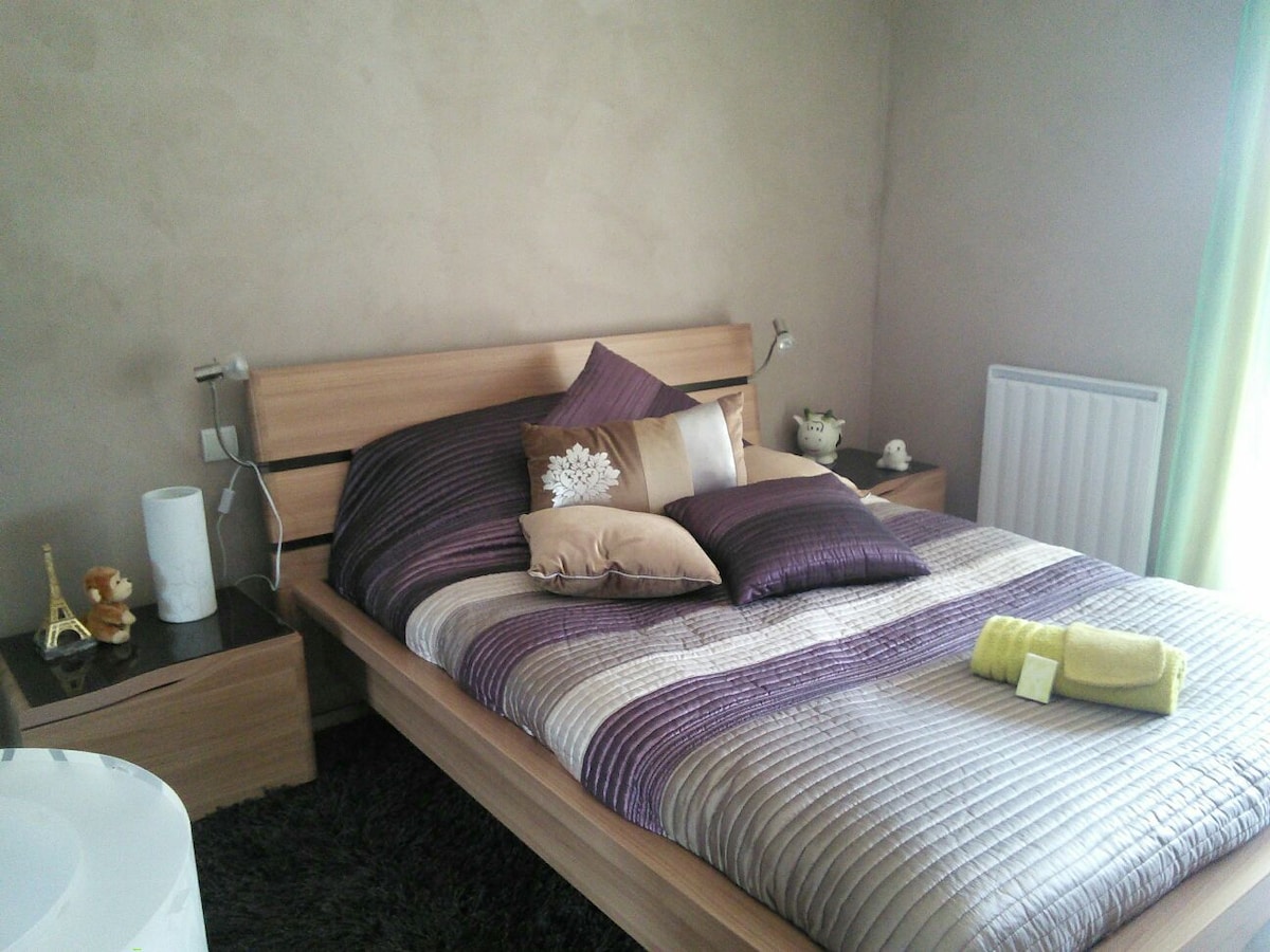 A sleeping area features a wooden bed with a quilted cover in shades of purple and grey. Decorative pillows add texture. A bedside table holds a teapot and a small decorative item. Natural light streams through curtains, illuminating the warm wall color.