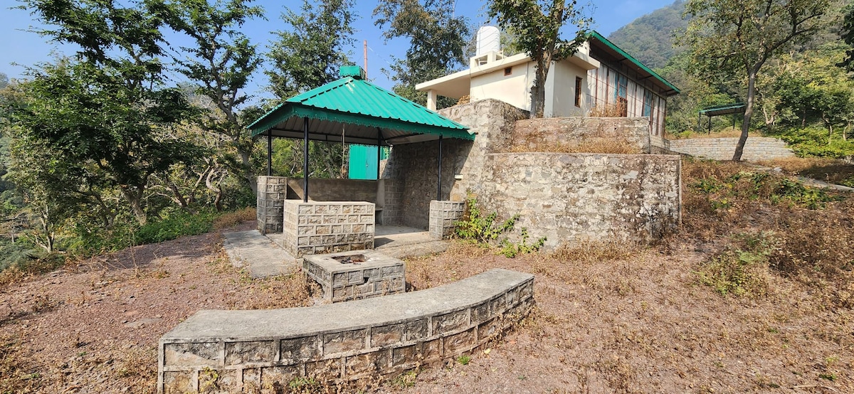 A stone-built structure is complemented by a green-roofed gazebo, providing a shaded area for relaxation. Surrounding greenery and hills create a serene environment. The pathway leads up to the building, with steps and natural landscaping visible in the area.