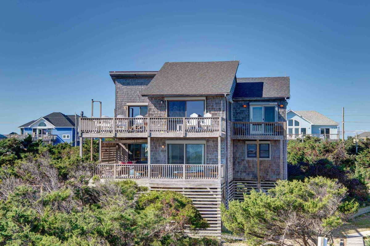 A modern oceanfront cottage is prominently displayed, featuring multiple decks and large windows. Surrounding greenery adds a natural touch, while neighboring homes are visible in the background. The inviting structure showcases a combination of wood and contemporary design elements.