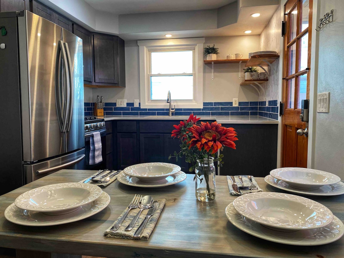 A newly renovated kitchen is highlighted by stainless steel appliances and dark cabinetry. A dining table is set for four, featuring white plates, silverware, and a vase of colorful flowers. Large windows allow natural light to fill the space.