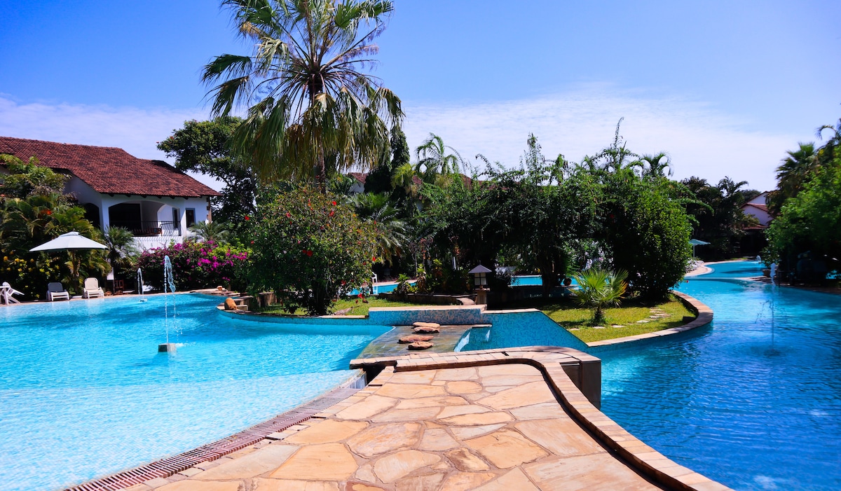 A three-tiered swimming pool is surrounded by lush greenery and tropical plants, offering a serene environment. The crystal blue water reflects the sky, while fountains add a gentle flow to the space. Lounge chairs are positioned near the pool for relaxation.