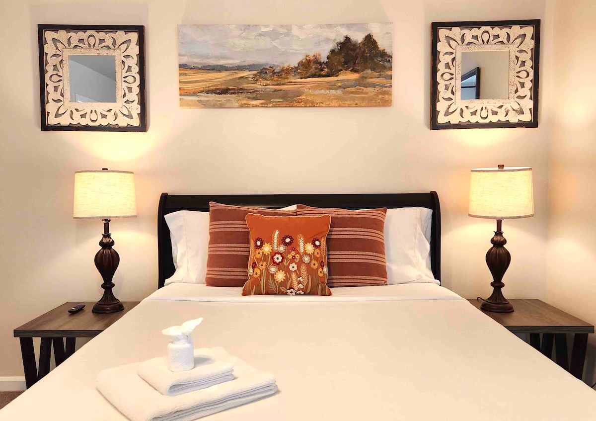 The room features a queen bed with a simple white duvet and decorative pillows. Two bedside tables with lamps are positioned on either side. Above the bed, two framed mirrors and a landscape painting add depth to the décor.