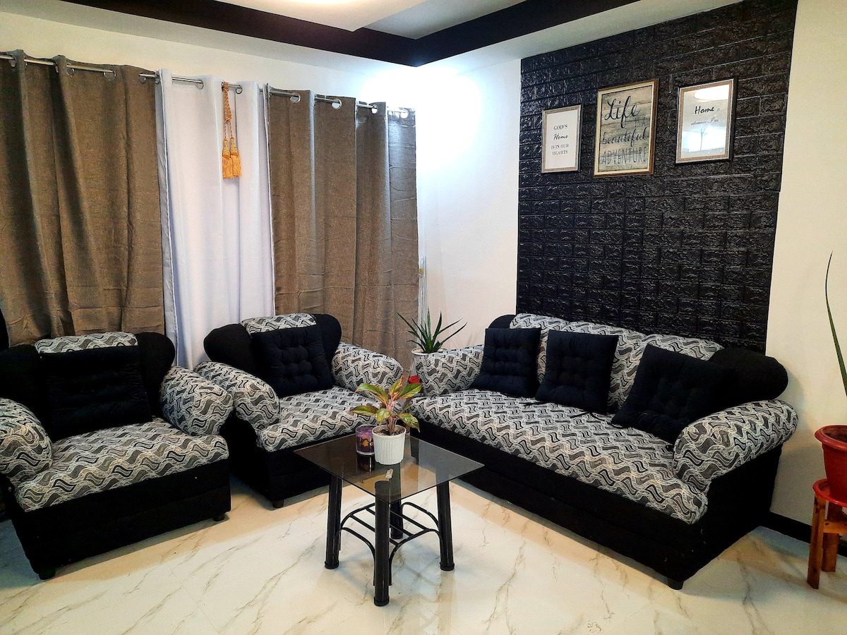 A comfortable living space features a pair of matching sofas adorned with patterned upholstery. A glass coffee table sits at the center, complemented by a small potted plant. Light flows through the sheer curtains, while darker drapes add depth to the room.