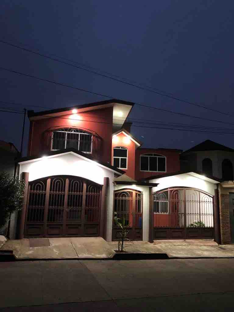 The exterior of a two-story home is illuminated by soft lighting at dusk. The building features a combination of red and cream-colored walls, with wrought iron gates and large windows that create a welcoming appearance.
