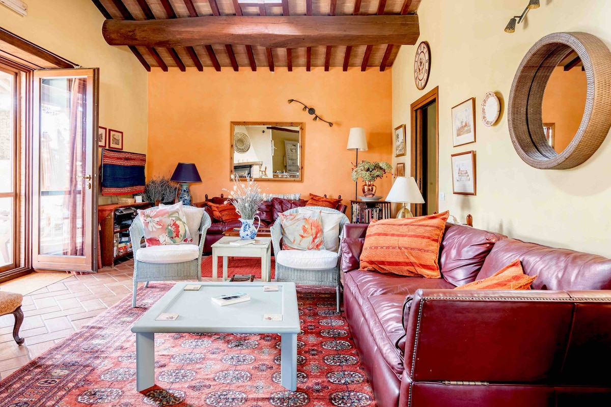 The living room features comfortable seating arrangements, with a deep red leather sofa and patterned armchairs. Bright orange walls complement wooden beams above. Natural light enters through open doors, creating a welcoming ambiance. A coffee table sits prominently, decorated with light-colored accents.