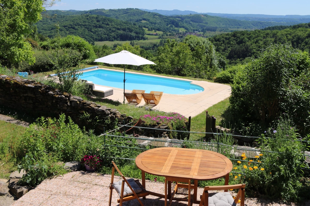 A spacious terrace is shown with a wooden dining table surrounded by chairs. In the background, a refreshing swimming pool is visible, bordered by lush greenery and a sun umbrella providing shade. Elevated views of the surrounding hills enhance the serene setting.