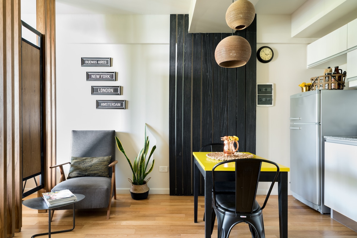 A modern living space features a sleek gray armchair beside a potted plant, with a dining table set for two. A yellow table adds a pop of color, complemented by hanging light fixtures. City names are displayed on the wall, contributing to the urban aesthetic.
