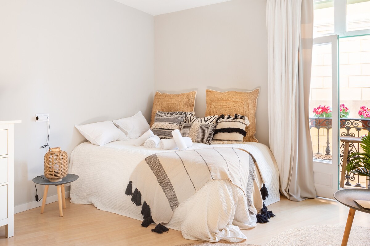 A spacious sleeping area is presented, featuring a textured blanket draped over a large bed with woven pillows. Natural light enters through balcony doors adorned with flowers. A small side table holds a decorative basket, while a plant adds a touch of greenery.