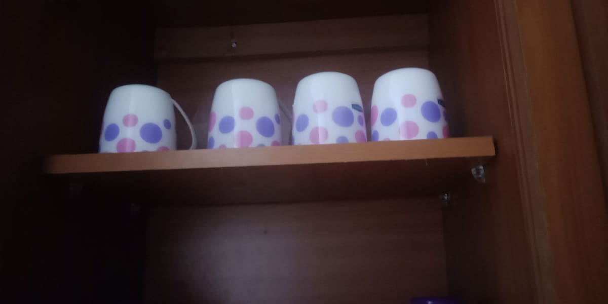 A wooden cabinet interior displays a shelf containing four white cups, each adorned with purple and pink polka dots. The simple design adds a playful touch to the organized space.