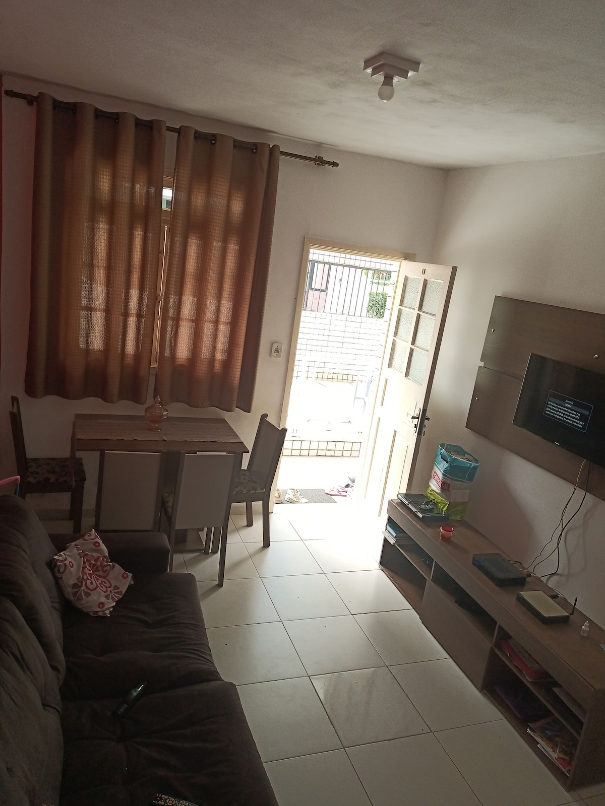 A living area features a cozy dark sofa and a small dining table with two chairs, complemented by warm brown curtains. Natural light filters through the door, which opens to an outdoor space. A wall-mounted TV and shelves are visible for entertainment.