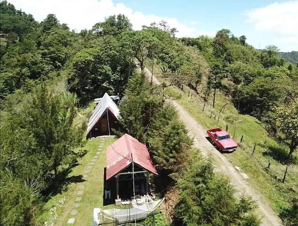 An aerial view captures a mountain chalet nestled among lush greenery. A dirt road leads to the property, with a vehicle parked nearby. The structure features a sloped roof and is surrounded by a neatly maintained lawn, creating a serene atmosphere in nature.