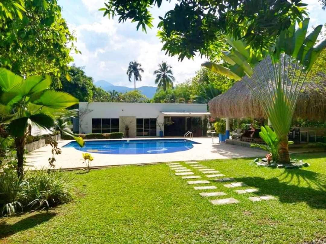 A private swimming pool is surrounded by lush greenery, with sun loungers placed along the water's edge. A thatched-roof hut is visible nearby, offering shaded seating. Pathways of stepping stones lead across the manicured lawn, providing access to different areas of the outdoor space.