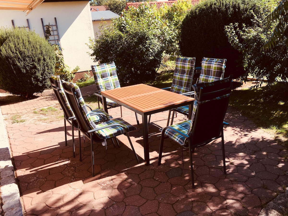 A patio area is presented with a rectangular table surrounded by six outdoor chairs. The chairs feature a plaid design, while the table has a wooden surface. Lush greenery and hedges provide a natural backdrop, enhancing the outdoor dining experience.