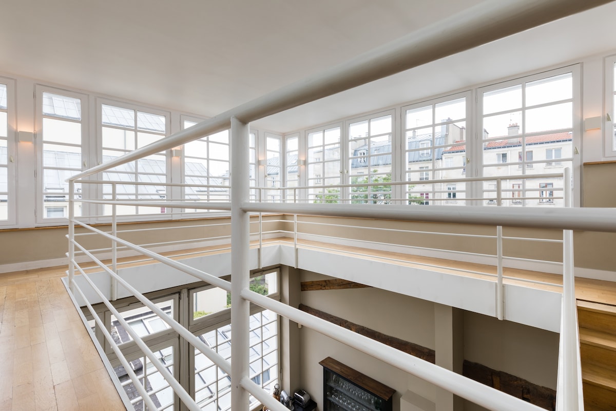 A spacious loft area is highlighted by an open design featuring a white railing. Large windows allow natural light to fill the space, offering views of the surrounding architecture. The warm wooden floors complement the airy atmosphere, enhancing the open feeling of the upper level.